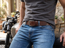 A close-up lifestyle shot of a man wearing a brown leather belt with a silver buckle, tucked into blue jeans, with a motorcycle in the background.