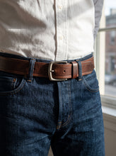 A close-up of a person wearing a brown leather belt with a silver-toned metal buckle, paired with dark blue jeans and a white button-up shirt, standing near a window.