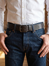 A close-up of a man wearing a dark brown embossed leather belt with a silver-toned metal buckle, paired with dark blue jeans and a white button-down shirt.