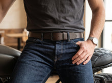 A close-up lifestyle shot of a man wearing a dark brown leather belt with a silver buckle, paired with dark blue jeans and a dark gray t-shirt, sitting on a black motorcycle seat.