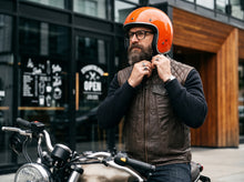A bearded man wearing a bright orange motorcycle helmet and a brown quilted leather vest over a black long-sleeve shirt, standing next to a motorcycle in front of a modern building with large glass windows and wood paneling.
