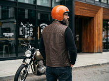 A man wearing a brown quilted leather vest, black long-sleeve shirt, and an orange helmet stands on a city sidewalk next to a motorcycle in front of a modern building with large glass windows and wood paneling.