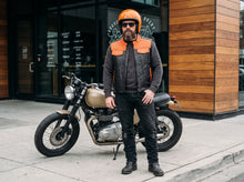 A bearded man wearing a black leather motorcycle jacket with bright orange quilted shoulder panels, a matching orange helmet, and black pants stands next to a cafe motorcycle on a city sidewalk.