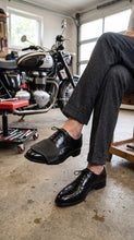 A person seated in a garage wearing black leather lace-up dress shoes and black leather motorcycle shifter boot protector