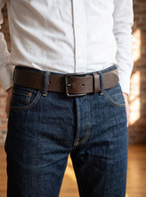 A man wearing a dark brown leather belt with a silver-toned rectangular buckle, tucked into dark blue denim jeans and paired with a white button-down shirt.