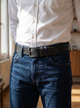 A close-up of a person wearing a dark textured leather belt with a silver-toned rectangular buckle, styled with a white button-down shirt and dark blue jeans in a brightly lit indoor setting.