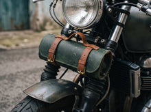 A dark green leather motorcycle saddle bag with brown leather straps and brass buckles, mounted on the front fork of a cruiser motorcycle.