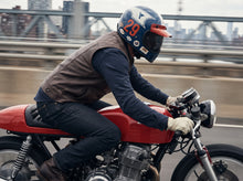 A person riding a red motorcycle on a bridge, wearing a blue, white, and orange full-face helmet, a brown leather vest, a dark long-sleeve shirt, and jeans.