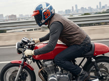 A person riding a red motorcycle on a highway, wearing a brown leather vest, dark jeans, gloves, and a blue racing helmet with the number 29. The background shows a blurred city skyline and guardrail.