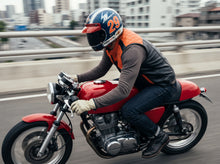 A person riding a red motorcycle on a city road, wearing a blue and orange full-face helmet, a black and orange vest, a brown long-sleeve shirt, and dark jeans.