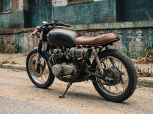 A custom cafe-style motorcycle parked on a street in front of a weathered brick building. The motorcycle features a matte black fuel tank, a brown leather seat with leather straps, a green leather side bag, and a chrome engine.