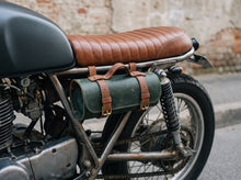 A close-up of a motorcycle with a brown quilted leather seat and a green cylindrical canvas bag strapped to the frame by brown leather straps, set against a blurred street background.