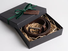 A dark brown leather belt with a silver buckle, presented inside a black gift box filled with shredded paper, tied with a green ribbon.
