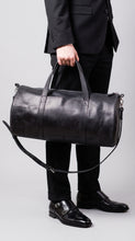 A person in a black suit holding a large black leather duffel bag with a shoulder strap against a light gray studio background.