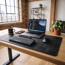 A large black leather desk mat on a wooden desk, styled with a mechanical keyboard, wireless mouse, laptop, notebook, and a coffee mug in a bright, modern office setting.