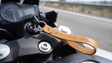 A close-up of a motorcycle keyring with a tan leather strap hanging near the handlebar. The strap is engraved with a phone number and the name 'Mark'.