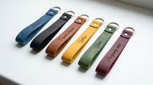 Five personalized leather keychains in different colors (blue, black, brown, yellow, green, and burgundy) arranged diagonally on a white surface, each engraved with a name and featuring a silver metal keyring.