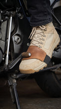 A close-up of a person's foot wearing a beige tactical motorcycle boot with Close-up of brown leather motorcycle shifter boot protector