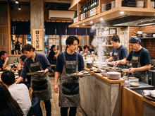 A group of restaurant chefs and waiters in matching navy blue shirts and gray aprons serving food in a warm, rustic Japanese-style dining space with visible steam rising from dishes.