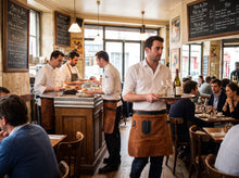 A restaurant interior with several waiters wearing white shirts and brown leather aprons serving tables. Patrons are seated at wooden tables under hanging lights, with chalkboard menus on the walls.