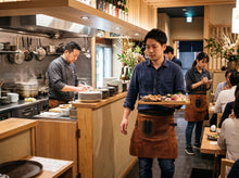A restaurant scene with a waiter in a brown leather apron carrying a tray of food, with kitchen staff and dining customers visible in a warm, wood-toned interior.