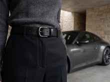 A close-up lifestyle shot of a person wearing a black leather belt with a silver-tone metal buckle, paired with dark trousers and a gray sweater. The background shows a gray sports car parked inside a stone-lined garage.