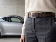 A close-up of a person wearing a dark brown leather belt with geometric orange stitching and a silver buckle, paired with gray trousers and a cream sweater. A silver sports car is blurred in the background.