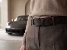 A close-up lifestyle shot of a person wearing a dark brown leather belt with a silver buckle and geometric stitched details, paired with a beige sweater and brown trousers, with a blurred Porsche in the background.