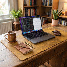 A styled home office desk setup featuring a yellow leather desk mat with a laptop, notebook, pens, and mug, set on a rustic wooden desk with a bookshelf and plants in the background.