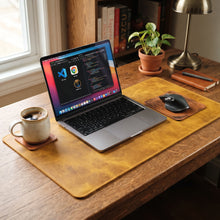 A large mustard-yellow leather desk mat on a wooden desk, styled with a laptop, mouse, mug, and a small potted plant in a home office setting.