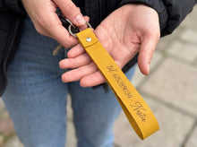 A close-up of a person holding a yellow leather keychain with a metal key ring and keys. The keychain has a snap button closure and is engraved with the text 'tel 615289034 Nattä'.