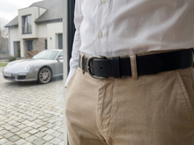 Black leather belt with rounded metal buckle styled with beige trousers and white shirt.