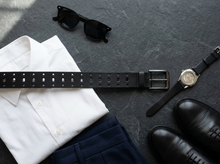 Black perforated leather belt styled with white shirt, navy trousers, watch, and sunglasses