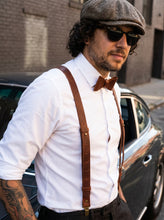 Man wearing a white shirt, brown suspenders, bow tie, and cap with sunglasses, standing next to a car.