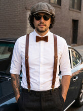 Man wearing brown leather suspenders, bow tie, and cap standing next to a car.