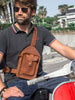 Man wearing a black polo shirt and blue jeans, sitting on a red scooter, with a brown leather crossbody bag featuring detailed stitching and multiple pockets. Handcrafted men's brown leather crossbody bag with detailed stitching, adjustable buckle and strap, and multiple pockets. Perfect for stylish and functional everyday use.