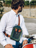 Man wearing a white shirt and blue jeans, with a handcrafted men's leather crossbody bag featuring green, brown, blue, and black leather, detailed stitching, and multiple pockets, sitting on a red scooter and looking to the side.