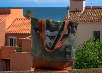 canvas tote bag with green, brown, and navy panels and leather straps, displayed against a scenic seaside rooftop view