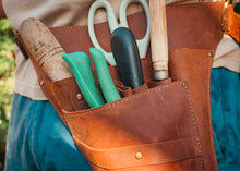 A close-up of a brown leather gardening tool belt worn by a person, filled with gardening tools including green-handled shears, a black-handled tool, and a wooden-handled implement.