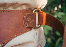 Close-up of a brown leather belt with brass hardware and a tan fabric strap, worn around the waist outdoors with blurred green foliage in the background.
