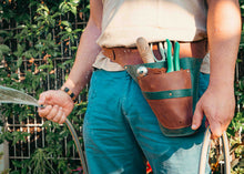 brown leather tool belt with gardening tools, holding a garden hose in a garden setting.
