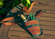 A brown leather gardening tool belt with green trim, shown on a wooden deck with gardening tools like shears and a hand fork tucked into its pockets.