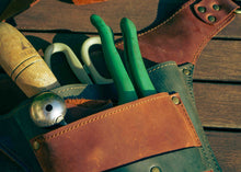 A close-up of a leather tool belt filled with hand tools, including green-handled pliers, white-handled shears, and a wooden-handled mallet, resting on a weathered wooden surface.