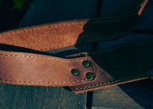A close-up of a brown leather strap with visible stitching and two metal rivets, laid on a dark wooden surface.