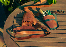 A brown leather gardening tool belt laid out on a wooden table, holding gardening tools including green-handled shears and a small hand tool. The belt has multiple pockets and a long strap,