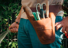 A brown leather gardening tool belt worn by a person, holding shears, a trowel, and other hand tools, with green foliage in the background.