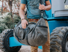 Person wearing a blue t-shirt and beige pants, holding a large gray duffel bag with black straps and a shoulder strap, standing next to industrial equipment with large tires.