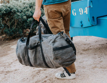 Man carrying a large, rugged gray waxed canvas duffel bag with black leather straps and a shoulder strap, wearing a blue shirt, beige pants, and white sneakers