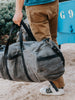 Man carrying a large, rugged gray waxed canvas duffel bag with black leather straps and a shoulder strap, wearing a blue shirt, beige pants, and white sneakers