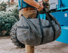 Person carrying a large, rugged gray duffel bag with black straps and a shoulder strap, standing next to industrial equipment.